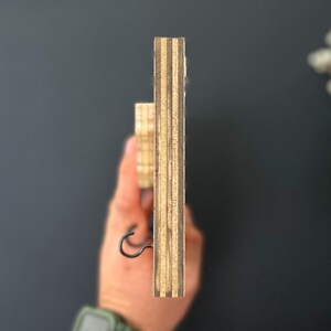 May include: Side view of a wooden key holder with a black metal hook. The key holder is made of plywood with visible layers of wood grain.