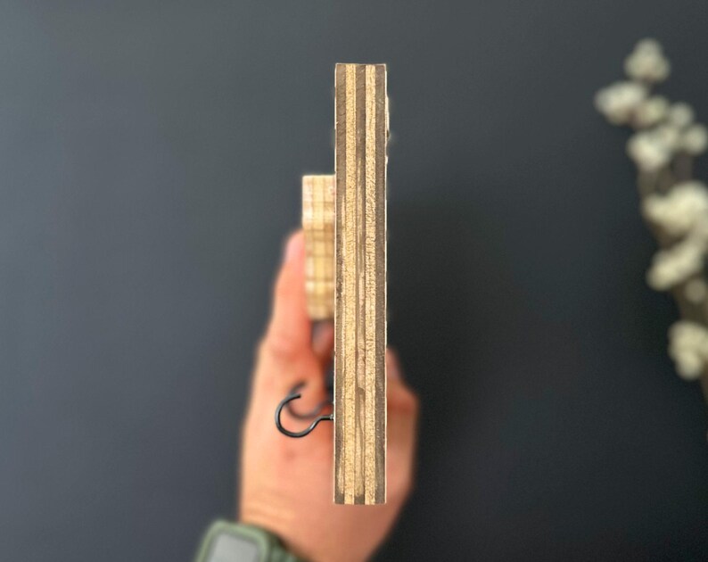 May include: Side view of a wooden key holder with a black metal hook. The key holder is made of plywood with visible layers of wood grain.