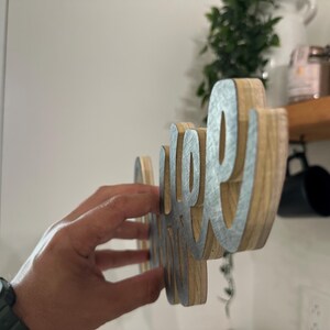May include: Wooden sign spelling "coffee" in a cursive font. The sign features a natural wood grain base with silver-coloured metal lettering. The sign is held up against a white wall, with a shelf and mugs visible in the background.