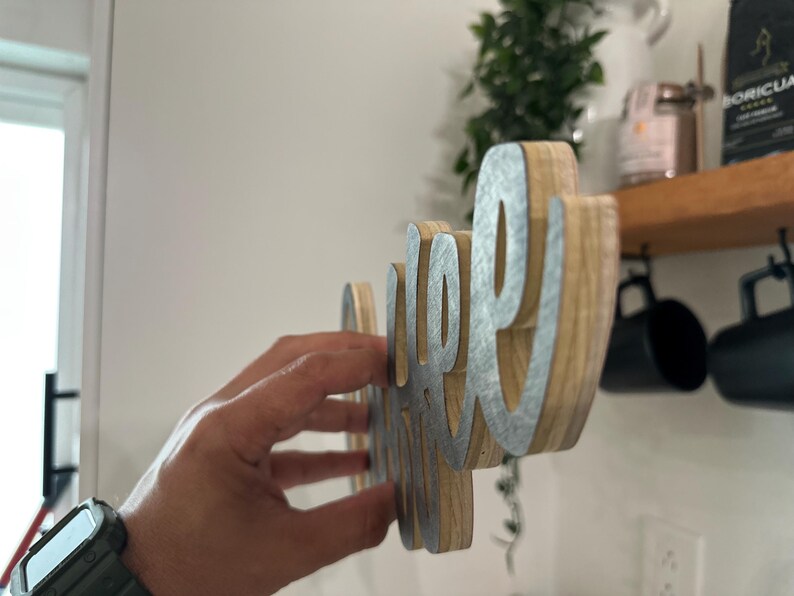 May include: Wooden sign spelling "coffee" in a cursive font. The sign features a natural wood grain base with silver-coloured metal lettering. The sign is held up against a white wall, with a shelf and mugs visible in the background.