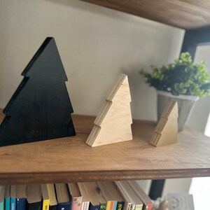 May include: Three wooden Christmas tree ornaments, one black and two natural wood. The ornaments are arranged on a wooden shelf with books below.
