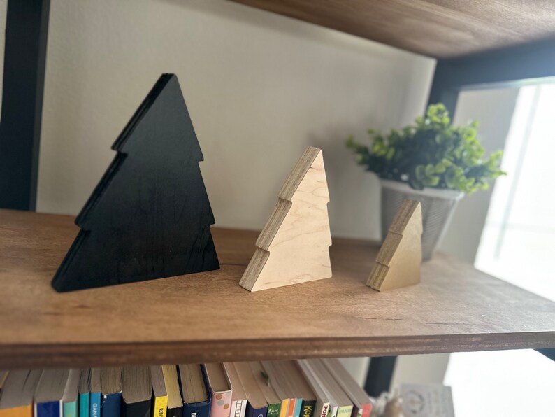 May include: Three wooden Christmas tree ornaments, one black and two natural wood. The ornaments are arranged on a wooden shelf with books below.