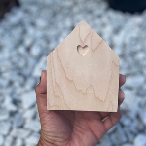 May include: A small wooden house shaped ornament with a heart cutout. The house is light brown and has a natural wood grain.