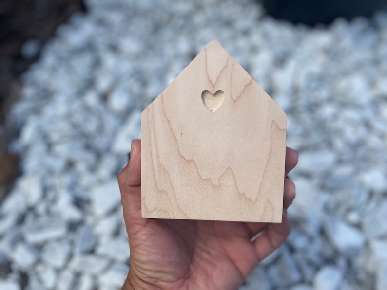 May include: A small wooden house shaped ornament with a heart cutout. The house is light brown and has a natural wood grain.