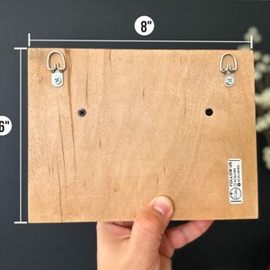 May include: The back of a wooden sign with two metal hanging hooks, measuring 20.3 cm wide by 15.2 cm tall. The sign has a small sticker that says "Follow Us" with a website address.