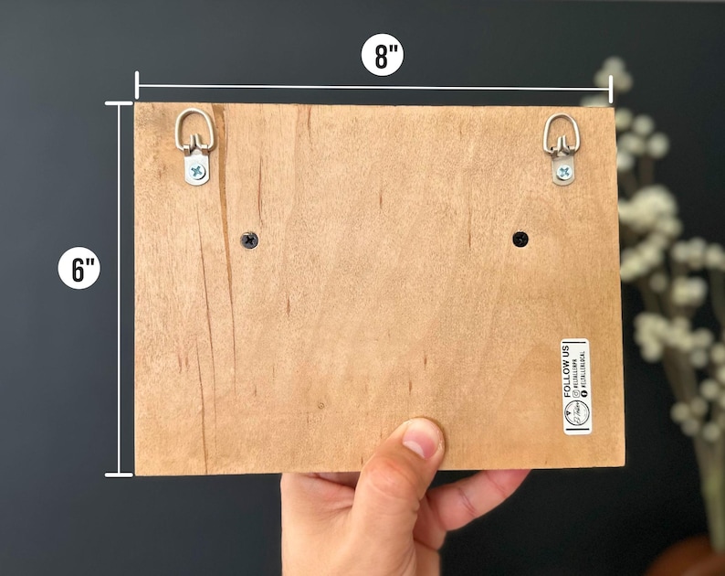 May include: The back of a wooden sign with two metal hanging hooks, measuring 20.3 cm wide by 15.2 cm tall. The sign has a small sticker that says "Follow Us" with a website address.