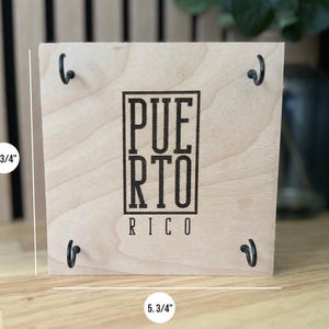 K&ouml;nnte beinhalten: Ein quadratisches Holzschild, 14,6 cm pro Seite, mit dem Wort "PUERTO RICO" in schwarzen Blockbuchstaben. Das Schild hat vier schwarze Metallhaken und eine nat&uuml;rliche Holzmaserung.