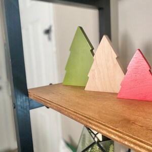 May include: Three wooden Christmas tree ornaments in different colors: green, natural wood, and red. The ornaments are arranged on a wooden shelf.