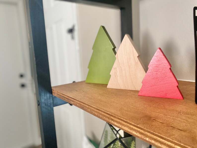 May include: Three wooden Christmas tree ornaments in different colors: green, natural wood, and red. The ornaments are arranged on a wooden shelf.