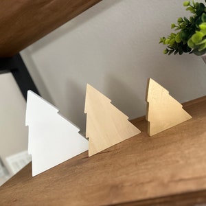 May include: Three wooden Christmas tree ornaments in different sizes. The largest ornament is white, the middle ornament is natural wood, and the smallest ornament is gold.