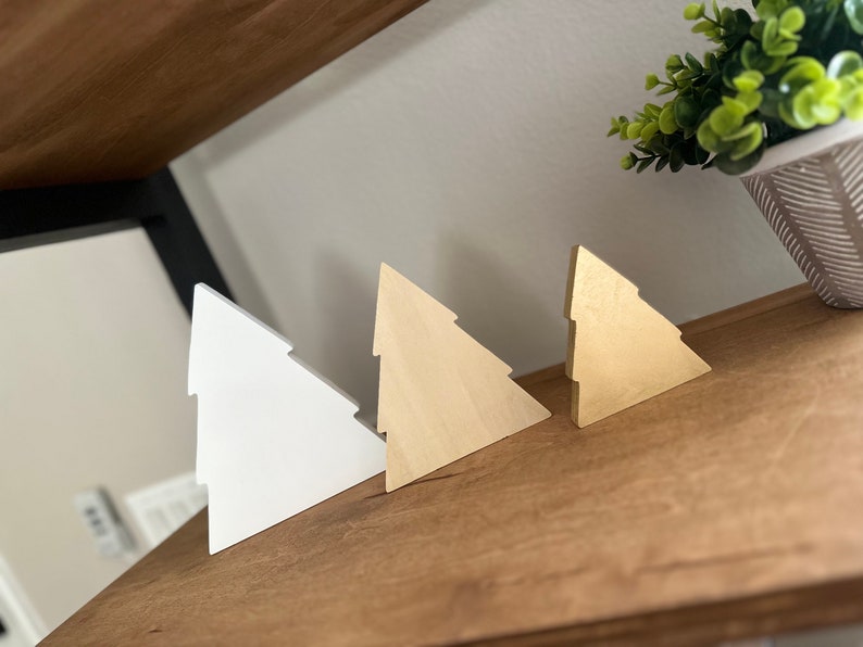 May include: Three wooden Christmas tree ornaments in different sizes. The largest ornament is white, the middle ornament is natural wood, and the smallest ornament is gold.