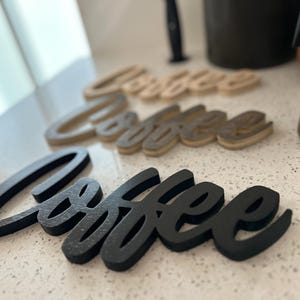 Pu&ograve; includere: Tre insegne in legno "Coffee" in varie tonalit&agrave; di marrone e nero sono disposte su una superficie bianca maculata. Le insegne sono in un carattere corsivo. Sullo sfondo c'&egrave; una caffettiera in acciaio inossidabile.