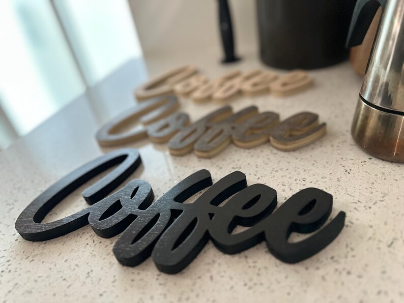 Pu&ograve; includere: Tre insegne in legno "Coffee" in varie tonalit&agrave; di marrone e nero sono disposte su una superficie bianca maculata. Le insegne sono in un carattere corsivo. Sullo sfondo c'&egrave; una caffettiera in acciaio inossidabile.