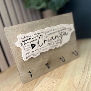 May include: Wooden key holder with a light brown finish. A white cutout in the shape of Puerto Rico is affixed to the wood, with the word "Crianza" in cursive. The key holder has four black hooks.