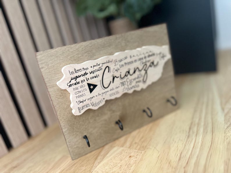 May include: Wooden key holder with a light brown finish. A white cutout in the shape of Puerto Rico is affixed to the wood, with the word "Crianza" in cursive. The key holder has four black hooks.