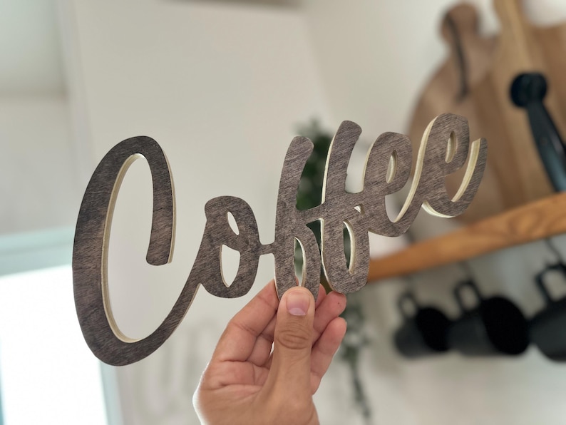 May include: A wooden sign spelling "Coffee" in a cursive font. The sign is dark brown with a lighter brown border. The sign is held up in front of a kitchen setting.
