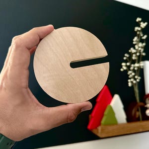 Puede incluir: Un posavasos redondo de madera con un corte en forma de letra "C". El posavasos es de color marr&oacute;n claro y tiene una veta de madera natural.