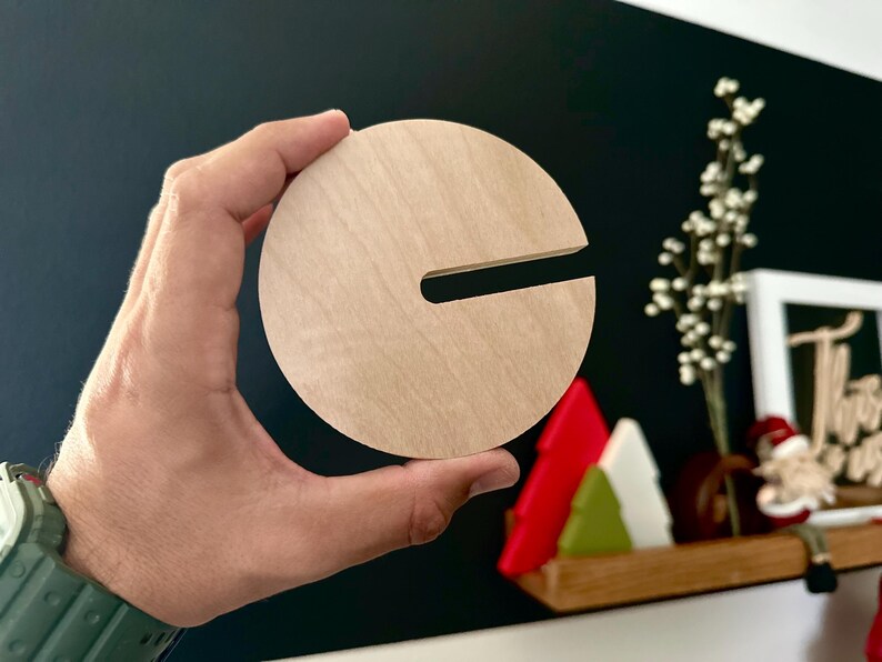 Puede incluir: Un posavasos redondo de madera con un corte en forma de letra "C". El posavasos es de color marr&oacute;n claro y tiene una veta de madera natural.