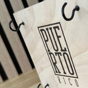 K&ouml;nnte beinhalten: Ein Holzschild mit dem Schriftzug "PUERTO RICO" in schwarzer, fetter Schrift. Das Schild hat schwarze Metallhaken und ist vor einem Hintergrund aus schwarzen und beigen vertikalen Streifen angebracht. Das Schild ist aus hellem Holz gefertigt.