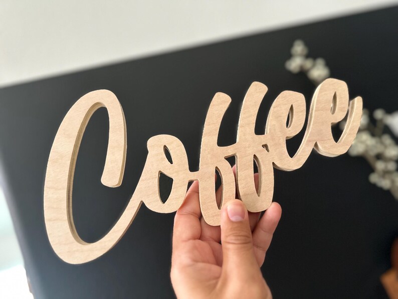 Pu&ograve; includere: Un cartello in legno con la parola "Coffee" scritta in corsivo. Il cartello &egrave; in legno di colore chiaro e viene tenuto contro uno sfondo scuro. &Egrave; un elemento decorativo per una cucina o un angolo caff&egrave;.