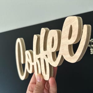 Pu&ograve; includere: Un cartello in legno con la scritta "Coffee" in corsivo. Il cartello &egrave; realizzato in legno chiaro e ha un effetto tridimensionale. Lo sfondo &egrave; un mix di nero e bianco.