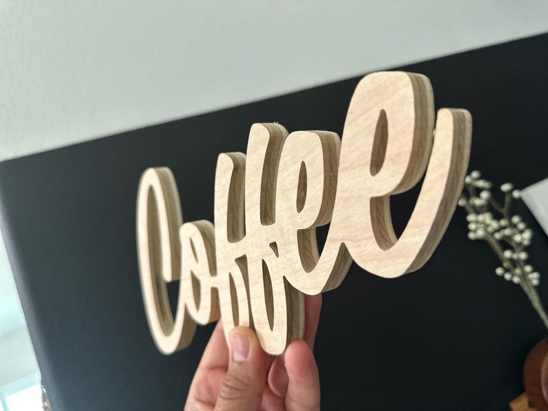 Pu&ograve; includere: Un cartello in legno con la scritta "Coffee" in corsivo. Il cartello &egrave; realizzato in legno chiaro e ha un effetto tridimensionale. Lo sfondo &egrave; un mix di nero e bianco.
