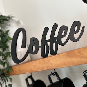 May include: A black wooden sign spelling "Coffee" in a cursive font. The sign is on a wooden shelf, with black mugs hanging beneath. Green foliage is visible on the left side of the image.