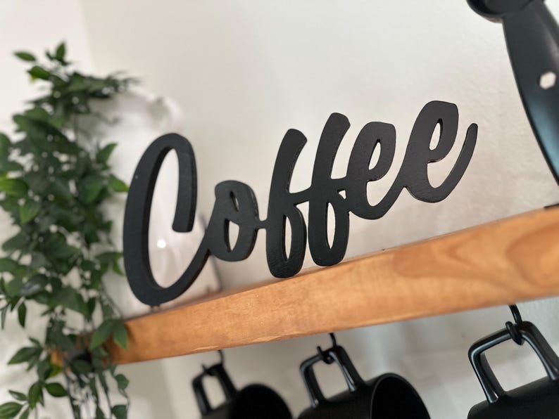 May include: A black wooden sign spelling "Coffee" in a cursive font. The sign is on a wooden shelf, with black mugs hanging beneath. Green foliage is visible on the left side of the image.
