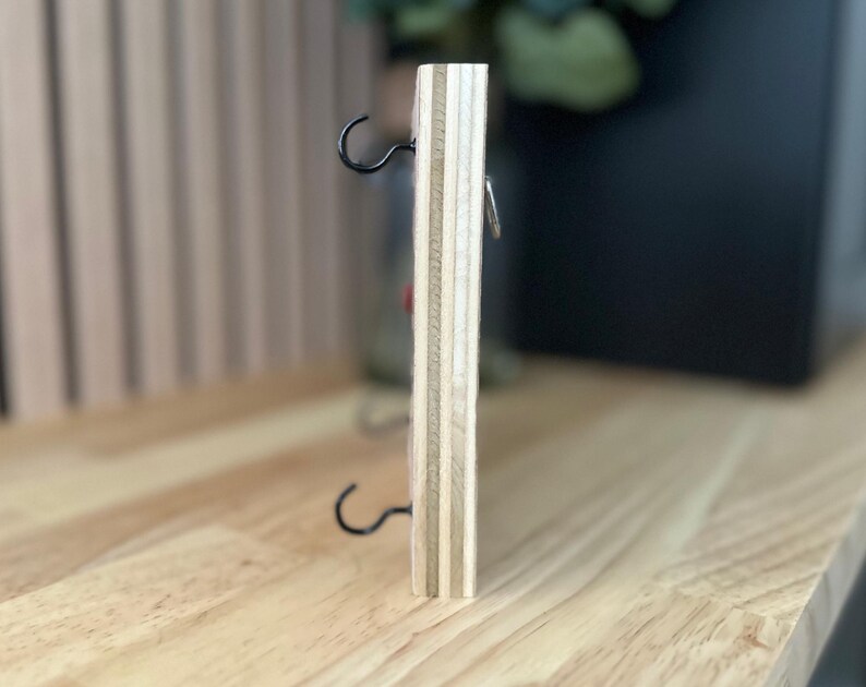 May include: A wooden wall-mounted key holder with two metal hooks and a small metal ring. The key holder is made of light wood with a natural finish.