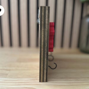 K&ouml;nnte beinhalten: Ein Wand-Schl&uuml;sselhalter aus Holz mit einem roten Blumendesign und zwei Metallhaken.