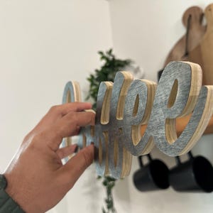 May include: A layered wooden sign spelling "coffee". The letters have a grey metallic front and a natural wood edge. The sign is being held up, with a shelf and mugs in the background.