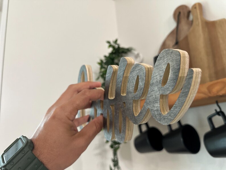 May include: A layered wooden sign spelling "coffee". The letters have a grey metallic front and a natural wood edge. The sign is being held up, with a shelf and mugs in the background.
