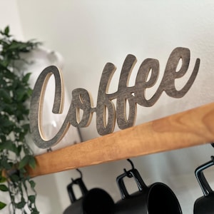 May include: Wooden "Coffee" sign in a cursive font, mounted on a wooden shelf. The sign is a light brown colour with a darker outline. Below the shelf are black coffee mugs. Green leafy plant in the background.