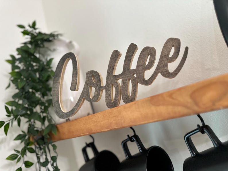 May include: Wooden "Coffee" sign in a cursive font, mounted on a wooden shelf. The sign is a light brown colour with a darker outline. Below the shelf are black coffee mugs. Green leafy plant in the background.