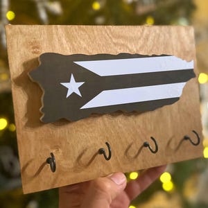 May include: Wooden key holder shaped like the island of Puerto Rico with the Puerto Rican flag painted on it. The key holder has five black metal hooks.