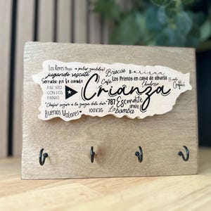 May include: A wooden key holder with a light finish. It has a cutout in the shape of Puerto Rico with the word "Crianza" and other text. Four black hooks are attached to the bottom of the wooden board, which measures 100 x 35 mm.