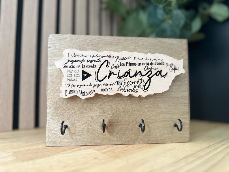 May include: A wooden key holder with a light finish. It has a cutout in the shape of Puerto Rico with the word "Crianza" and other text. Four black hooks are attached to the bottom of the wooden board, which measures 100 x 35 mm.