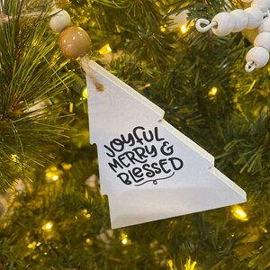 May include: White wooden Christmas tree bauble with the text "Joyful Merry & Blessed" in black lettering.