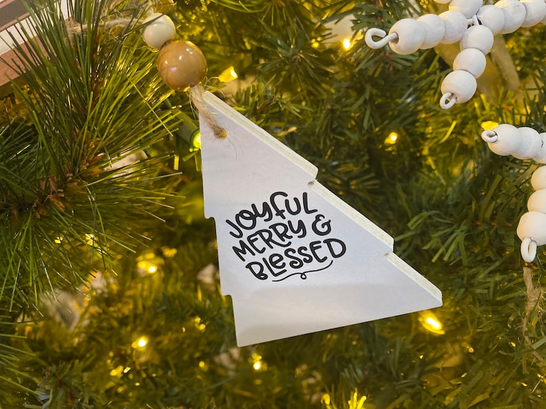 May include: White wooden Christmas tree bauble with the text "Joyful Merry & Blessed" in black lettering.