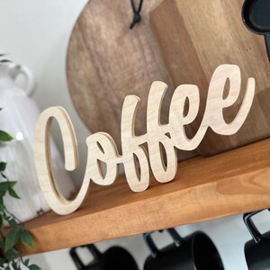 Pu&ograve; includere: Un'insegna in legno chiaro con la scritta "Coffee" in corsivo. L'insegna &egrave; posta su una mensola di legno. Sotto, sono appese delle tazze nere, con un vaso bianco e del verde sullo sfondo.