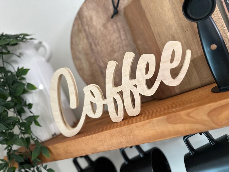 Pu&ograve; includere: Un'insegna in legno chiaro con la scritta "Coffee" in corsivo. L'insegna &egrave; posta su una mensola di legno. Sotto, sono appese delle tazze nere, con un vaso bianco e del verde sullo sfondo.