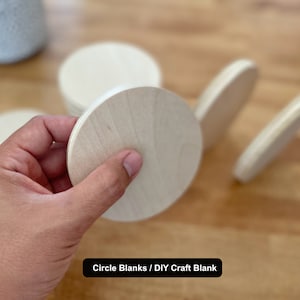 May include: A stack of round wooden craft blanks, also known as circle blanks, are shown on a light wood surface. A hand is holding one of the blanks. The text "Circle Blanks / DIY Craft Blank" is visible at the bottom of the image.
