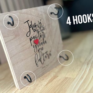 May include: A wooden sign with the text "Jehov&aacute; es mi pastor y nada me faltar&aacute;" in black cursive lettering with a red heart. The sign has four black metal hooks on the sides.