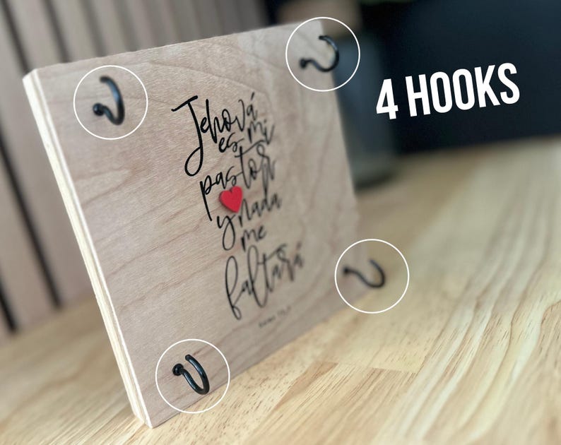 May include: A wooden sign with the text "Jehov&aacute; es mi pastor y nada me faltar&aacute;" in black cursive lettering with a red heart. The sign has four black metal hooks on the sides.