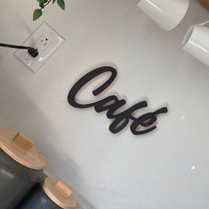 May include: Black wooden sign with the word "Cafe" written in a cursive font. The sign is mounted on a white wall.