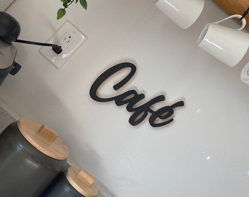 May include: Black wooden sign with the word "Cafe" written in a cursive font. The sign is mounted on a white wall.