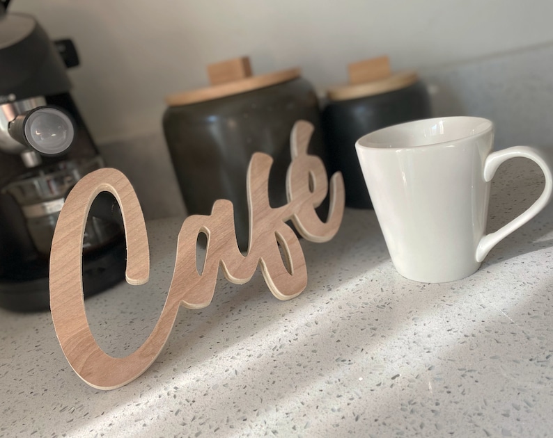 May include: A wooden sign that says "Cafe" in a cursive font. The sign is on a white countertop with a white coffee mug and a black coffee maker in the background.