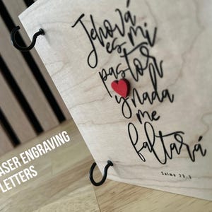 May include: Laser engraved wooden plaque with the text "Jehov&aacute; es mi pastor y nada me faltar&aacute;" in black cursive lettering. A small red heart is attached to the plaque. The text "Salmo 23:1" is also engraved on the plaque.