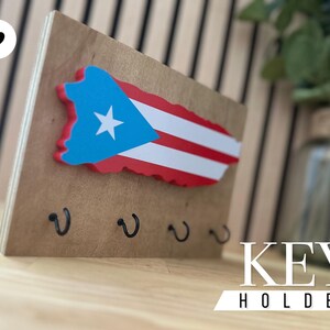 K&ouml;nnte beinhalten: Schl&uuml;sselhalter aus Holz in Form der Insel Puerto Rico mit der Flagge von Puerto Rico darauf bemalt. Der Schl&uuml;sselhalter hat vier Haken zum Aufh&auml;ngen von Schl&uuml;sseln. Der Text "KEY HOLDER" ist unter dem Schl&uuml;sselhalter aufgedruckt.
