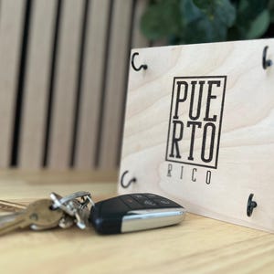 K&ouml;nnte beinhalten: Ein h&ouml;lzerner Schl&uuml;sselhalter mit dem schwarzen Aufdruck "PUERTO RICO". Der Schl&uuml;sselhalter hat vier schwarze Haken und wird von einem Schl&uuml;sselsatz und einer Auto-Fernbedienung begleitet. Der Schl&uuml;sselhalter ist aus hellem Holz gefertigt.